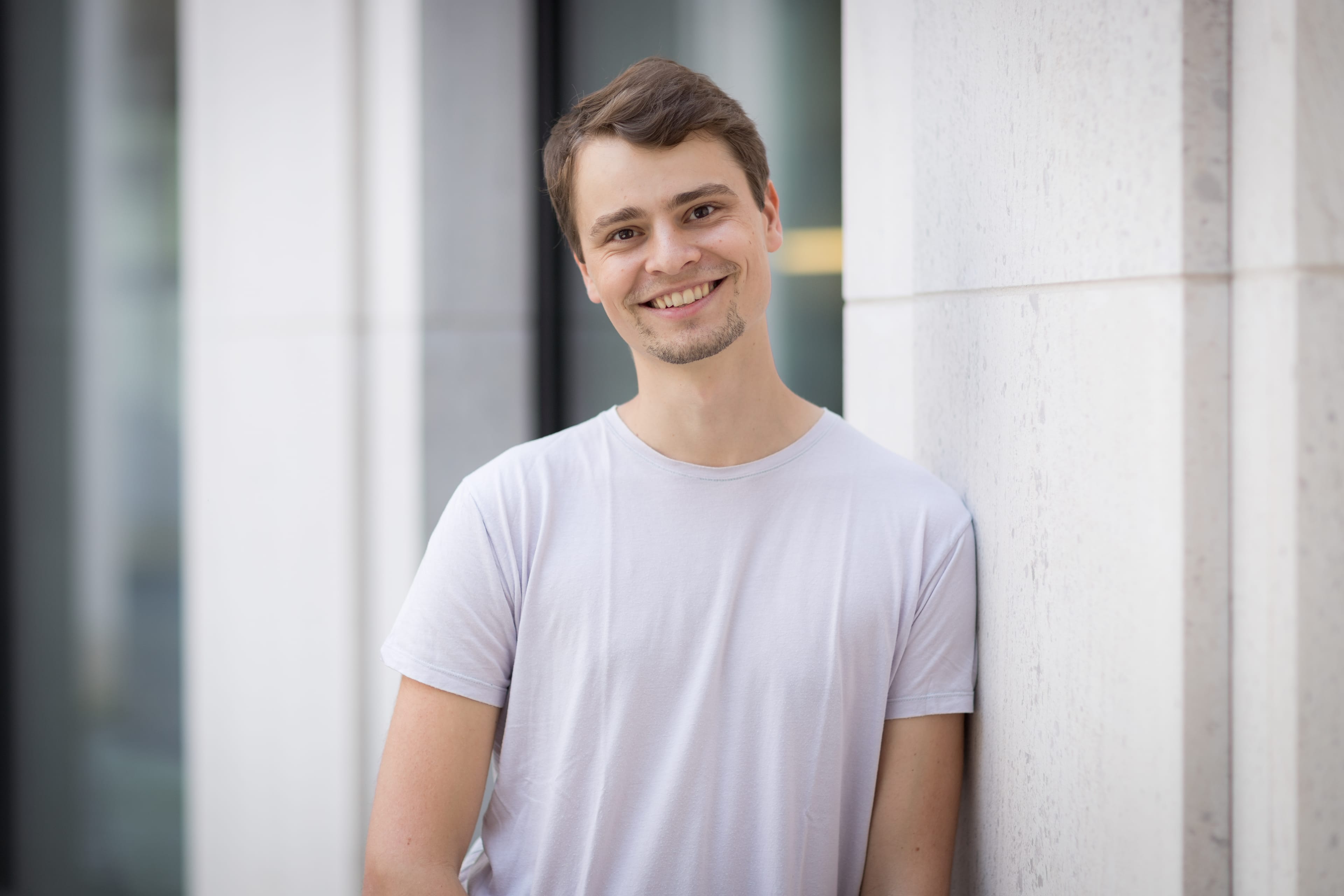 A young man leans against a modern office building. He has brown hair, a short extended goatee and wears a light lavender t-shirt.